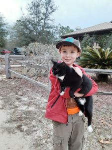 Young boy in ball cap and red hoodie holding black and white cat in front of camp house